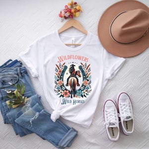 May include: White t-shirt with a graphic of a woman riding a horse with a horseshoe and flowers around it. The text "Wildflowers and Wild Horses" is printed on the shirt.