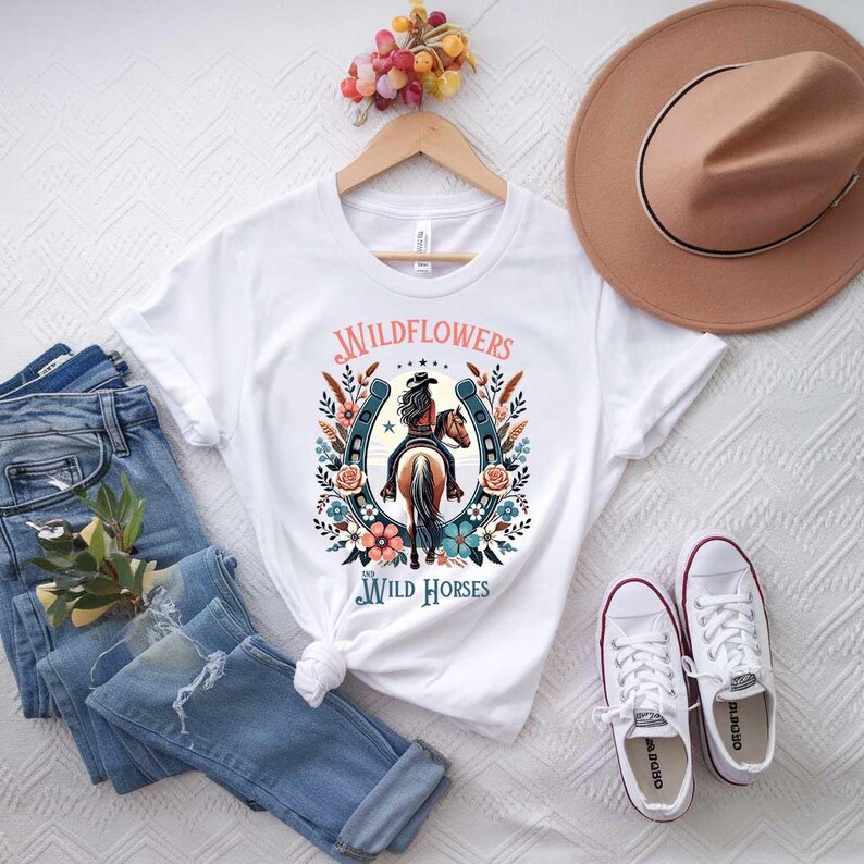 May include: White t-shirt with a graphic of a woman riding a horse with a horseshoe and flowers around it. The text "Wildflowers and Wild Horses" is printed on the shirt.