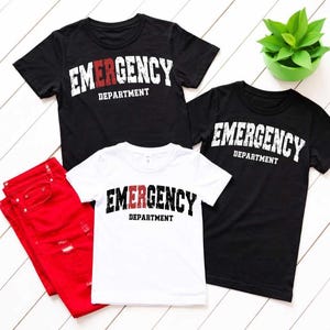 May include: Three t-shirts, two black and one white, with the text "EMERGENCY DEPARTMENT" in a bold, distressed font. The word "EMERGENCY" is white on the black shirts and black on the white shirt. The word "DEPARTMENT" is white on the black shirts and black on the white shirt. Red jeans are also in the image.