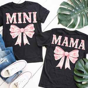 Puede incluir: Dos camisetas negras con un lazo floral rosa y las palabras "MINI" y "MAMA" impresas en ellas.