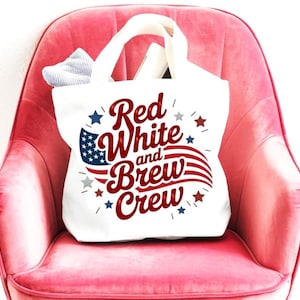 Könnte beinhalten: Eine weiße Stofftasche mit zwei Henkeln, die den rot-weiß-blauen Schriftzug "Red White and Brew Crew" in einer stilisierten Schriftart zeigt, begleitet von einer Grafik einer wehenden amerikanischen Flagge und mehreren kleinen roten, weißen und blauen Sternen.