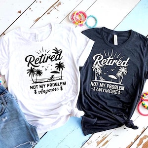 May include: Two t-shirts, one white and one dark gray, with the text "Retired, Not My Problem Anymore" and a beach scene graphic. The graphic includes palm trees, a beach chair, and an umbrella. The shirts are on a white wooden surface.