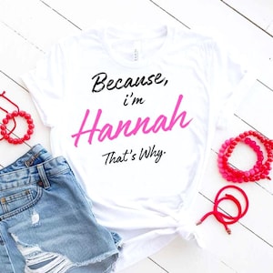 May include: White t-shirt with the text "Because, I'm Hannah That's Why" in black and pink lettering. The shirt is paired with denim shorts and red beaded bracelets on a white wooden surface.