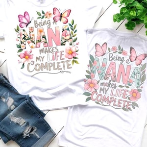 May include: Two white t-shirts with the text "Being a NANA makes my life COMPLETE" in a floral design. The word "NANA" is in pink and floral patterns. Pink butterflies and flowers surround the text. Blue denim jeans are folded below.