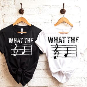 May include: Two t-shirts, one black and one white, each with the phrase "WHAT THE" above a musical staff with a treble clef and a quarter note. The shirts are hanging on wooden hangers against a white brick wall.