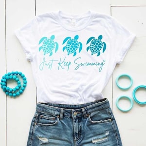 Puede incluir: Camiseta blanca con un gráfico de tres tortugas marinas azules y verdes y el texto "Just Keep Swimming".