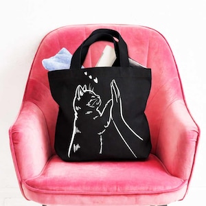 May include: Black tote bag with a white graphic of a cat high-fiving a hand. The cat is looking up with three small hearts above its head. The tote bag is sitting on a pink chair and contains a blue and white striped cloth.