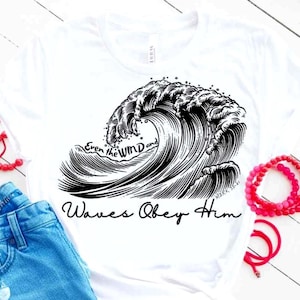 May include: White t-shirt featuring a black and white illustration of a large ocean wave. The text "Even the WIND and Waves Obey Him" is written in a script font. The shirt is paired with blue jeans and red bracelets.
