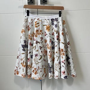 May include: A white A-line skirt with a floral pattern in shades of orange, purple, and pink. The skirt is hanging on a wooden hanger against a white background. The skirt has a white waistband.