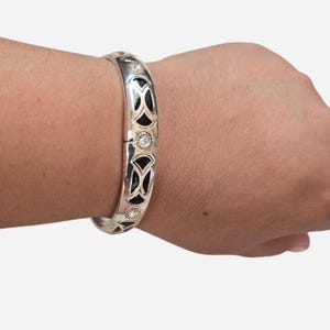 May include: Silver bangle bracelet with black enamel and clear crystal accents. The bracelet features a repeating pattern of black enamel shapes and small crystals. The bracelet is worn on a wrist.