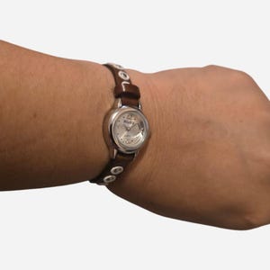 Könnte beinhalten: Eine silberfarbene Armbanduhr mit einem braunen Lederarmband. Das Zifferblatt der Uhr ist rund mit silbernen Zeigern und Stundenmarkierungen. Das Armband hat silberfarbene Nieten. Die Uhr ist von der Marke Mudd.