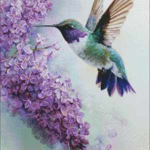 Puede incluir: Un patrón de punto de cruz que muestra un colibrí con plumas iridiscentes verdes, moradas y blancas volando cerca de un racimo de flores de lila de color morado intenso. El diseño está sobre un fondo de tela blanca con sombras suaves.