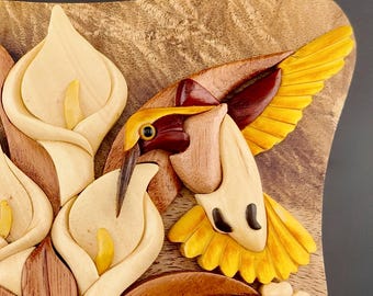 May include: A wooden hummingbird puzzle box with intricate details. The hummingbird is carved in shades of brown, red, and yellow, with its wings spread out. The box is decorated with carved white calla lilies.