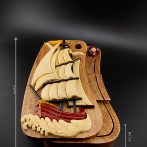 May include: A wooden nautical-themed box with a carved sailboat design. The box is 6.5 inches long, 4 inches wide, and 2.5 inches high. The sailboat has cream-colored sails and a red hull, with waves carved at the base.