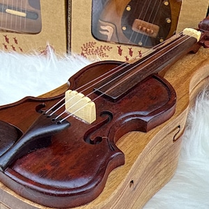 May include: A miniature wooden violin with a dark brown finish, resting on a light brown wooden case. The violin features strings, a bridge, and a scroll. The case has a curved design, and the overall aesthetic is rustic and handcrafted.