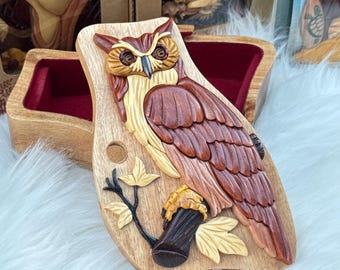 May include: A wooden owl carving with detailed feathers, perched on a branch with leaves. The owl is primarily brown, with lighter beige and cream accents. The carving is mounted on a wooden plaque, with a wooden box in the background.