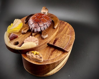 Puede incluir: Una caja de rompecabezas de madera con un diseño de tortuga marina tallada. La caja está hecha de madera oscura y tiene una tapa con bisagras. La tapa está decorada con una tortuga marina tallada, algas marinas y conchas.