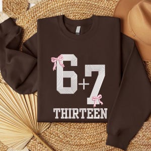 May include: A brown sweatshirt with the numbers "6 + 7" in white, with pink bow accents. The word "THIRTEEN" is printed in white below the numbers. The sweatshirt is displayed on a woven surface.