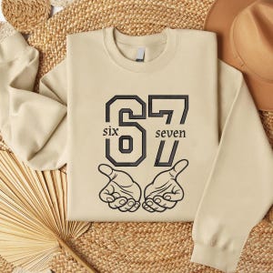 以下が含まれることがあります： ベージュ色のスウェットシャツ。黒色のカレッジ風フォントで「67」の数字がプリントされています。「six」の文字が6の下に、「seven」の文字が7の下にあります。数字の下には2つの手が描かれています。