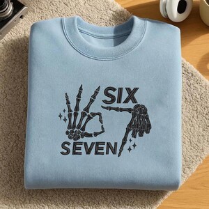 May include: A light blue sweatshirt with a crew neck. The front features a black graphic of two skeleton hands forming the number six and seven, with the words "SIX" and "SEVEN" printed below. The design includes star accents.