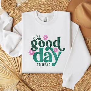 May include: White sweatshirt with the text "IT'S A GOOD DAY TO READ" in teal and green, with pink floral accents. The sweatshirt is displayed on a woven surface, with a brown hat visible in the background.