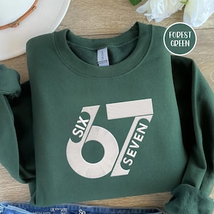 May include: Forest green sweatshirt with the number 67 in white block letters and the words "SIX" and "SEVEN" on either side. The sweatshirt is folded on a wooden surface, with a white hat and a flower in the background.
