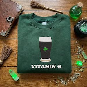 May include: A dark green sweatshirt with a graphic of a pint of stout beer with a shamrock and the text "VITAMIN G". The sweatshirt is surrounded by witchy items like a cauldron, crystals, and a book.