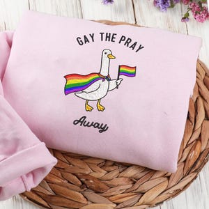 May include: A pale pink sweatshirt with a cartoon goose wearing a rainbow cape and holding a rainbow flag. The text "GAY THE PRAY AWAY" is arched above and below the goose illustration. The sweatshirt is folded on a woven surface.