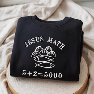 Puede incluir: Una sudadera negra con la inscripción blanca "JESUS MATH" y un dibujo de panes y peces. La ecuación "5+2=5000" está impresa en blanco debajo del dibujo.