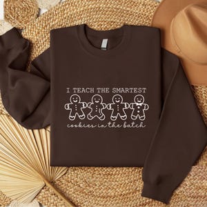 May include: A dark brown sweatshirt with the text "I teach the smartest cookies in the batch" and a graphic of four gingerbread men. The sweatshirt is made of a soft material and is perfect for teachers.
