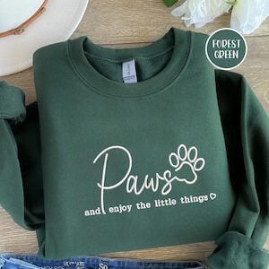 Puede incluir: Sudadera verde bosque con la palabra "Paws" en escritura cursiva y un gráfico de huella de pata. El texto "and enjoy the little things" está debajo de la palabra "Paws". La sudadera está doblada sobre una superficie de madera.