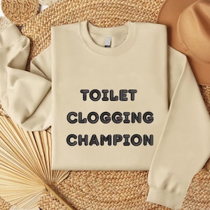 Puede incluir: Una sudadera beige con el texto "TOILET CLOGGING CHAMPION" en una fuente negra con burbujas. La sudadera se muestra sobre una superficie tejida con un sombrero marrón y un abanico de hojas de palma secas en el fondo.
