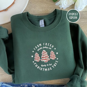 Puede incluir: Sudadera verde bosque con cuello redondo. La parte delantera presenta un gráfico de tres árboles de Navidad con glaseado rojo y blanco, y el texto "FARM FRESH Sprinkles frosting cake CHRISTMAS TREE Ready to Eat".