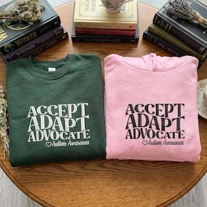 May include: Two sweatshirts, one forest green and one pink, each with the words "ACCEPT, ADAPT, ADVOCATE" in white and black, respectively. Below the text is "Autism Awareness" with a heart symbol. The sweatshirts are on a wooden table.
