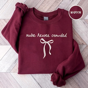 May include: Maroon sweatshirt with the phrase "make heaven crowded" in white script, accompanied by a white bow graphic. The sweatshirt is folded, showcasing the design. A circular label in the upper right corner reads "MAROON".