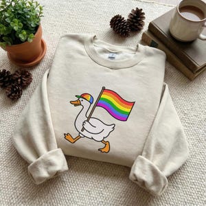 May include: Cream-colored sweatshirt featuring a cartoon goose wearing a rainbow cap and carrying a rainbow flag. The goose is running, with orange feet and a yellow beak. The design is centered on the front of the long-sleeved crewneck.