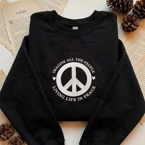 Può includere: Felpa girocollo nera con un simbolo della pace bianco e la scritta "IMAGINE ALL THE PEOPLE LIVING LIFE IN PEACE". Il capo è piegato su una superficie con pagine di libro e pigne.