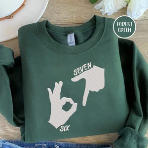 May include: Forest green sweatshirt with the words "SEVEN SIX" and hand gestures in white. The sweatshirt is folded on a wooden surface. A white circle with the words "FOREST GREEN" is in the upper right corner.