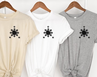 May include: Three t-shirts with a black sun and star design. The shirts are beige, white, and gray.