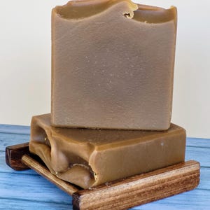 May include: Two bars of brown handmade soap on a wooden soap dish. The soap has a textured surface and an uneven top. The wooden dish is rectangular with a slatted design. The soap dish is made of wood.