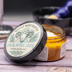 Puede incluir: Un tarro de jabón de afeitar artesanal con la tapa quitada, que revela una sustancia amarilla y blanca. La tapa es negra con las palabras "Woolly Wash Soapery" y "Eucalyptus Chill". Una brocha de afeitar y una navaja están en el fondo.
