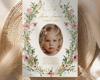 May include: A floral-themed first birthday invitation with a photo of a young girl in a white dress and a pink bow. The invitation features a wreath of pink and white flowers, green leaves, and vines. The text on the invitation reads "Ava Jane Turns One" and includes the date, time, address, and RSVP information.