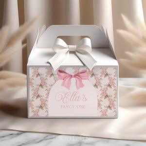 May include: White favour box with a pink floral pattern and a pink bow. The box has a white ribbon and bow on top. The text on the box reads "Ella's Fancy One".