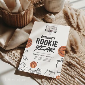 May include: A white invitation with black and orange basketball-themed illustrations. The text reads "Dominic's Rookie Year" and includes details for a first birthday party.