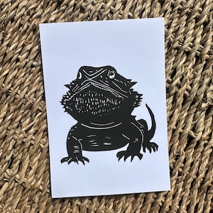 May include: A black and white linocut print of a bearded dragon on a white card. The reptile is depicted in a detailed, stylized manner, with spiky features and a textured beard. The card is set against a woven, natural-toned background.