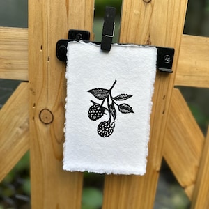 May include: A rectangular piece of white paper with a deckled edge featuring a black ink block print of two raspberries on a stem with leaves. The paper is held by a black clip attached to a wooden structure, displaying a botanical illustration.