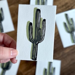 May include: A hand holds a small white card featuring a black and green cactus illustration. The cactus has a dark green shadow, and the image is repeated on other cards in the background. The cards are on a wooden surface.