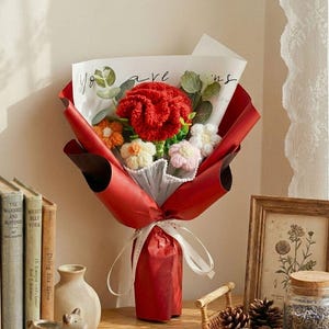 May include: A bouquet of handmade crochet flowers wrapped in red paper, tied with a white ribbon. The bouquet includes a large red flower, and smaller orange, white, and pink flowers. The bouquet is on a wooden surface with books and decorative items.
