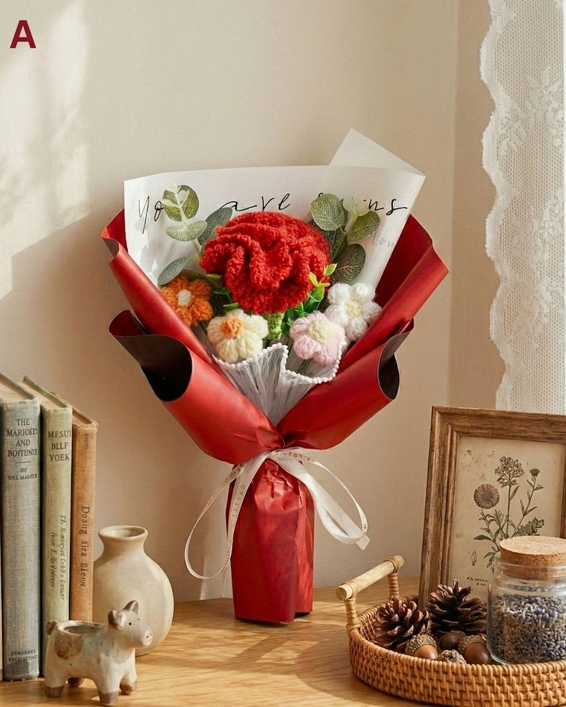 May include: A bouquet of handmade crochet flowers wrapped in red paper, tied with a white ribbon. The bouquet includes a large red flower, and smaller orange, white, and pink flowers. The bouquet is on a wooden surface with books and decorative items.