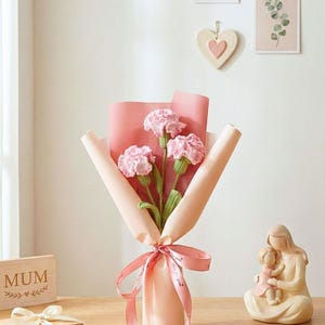 May include: A bouquet of pink crocheted carnations wrapped in pink paper and tied with a pink ribbon. The word "Pink" is in the top left corner. A small gift box and a wooden sign that says "MUM" are also visible.